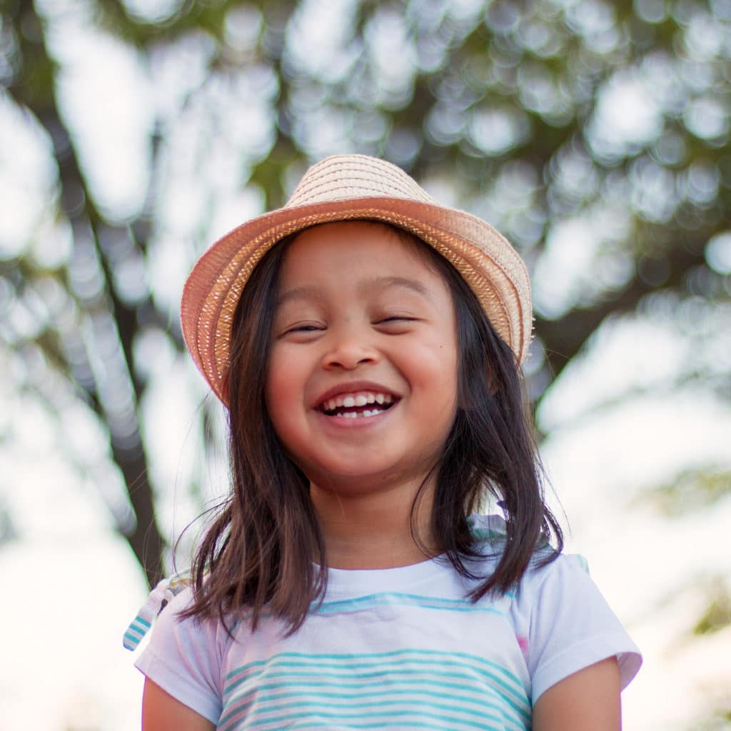 Little girl wearing a hat smiling in front of a tree