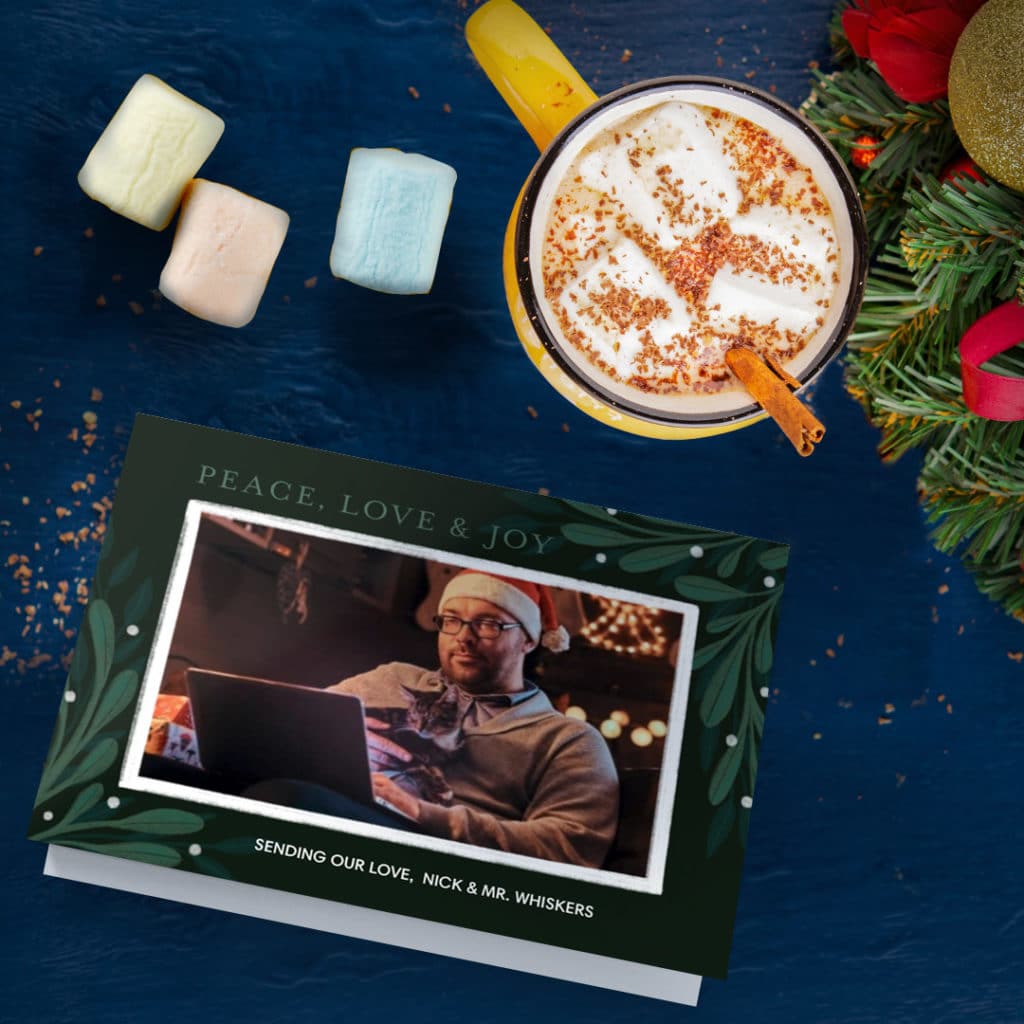 greeting card featuring wan cuddling with cat in front f laptop, surrounded by hot cocoa and marshmallows