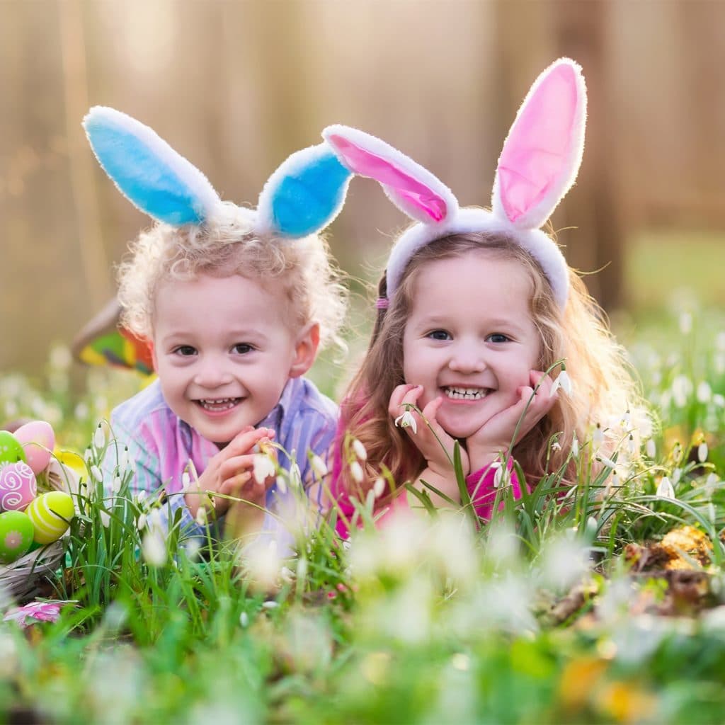 Two toddlers laying in the grass, surrounded by easter eggs, wearing pink and blue bunny ears.