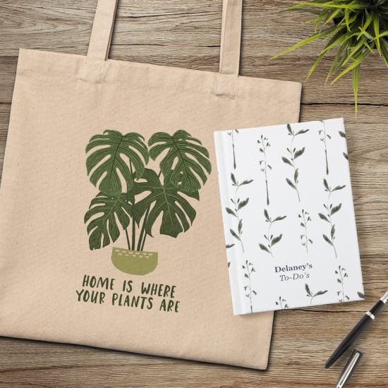 Canvas tote bag featuring "Home is where your plants are" design laying with a hardcover notebook featuring a leafy greenery design