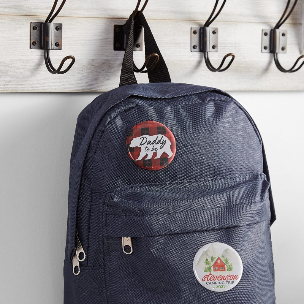 Daddy-to-be and "Camping Trip" pins shown on a navy blue backpack hanging from a hook