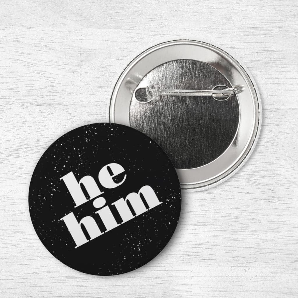 Black pin laying on a table with a design reading "He Him"