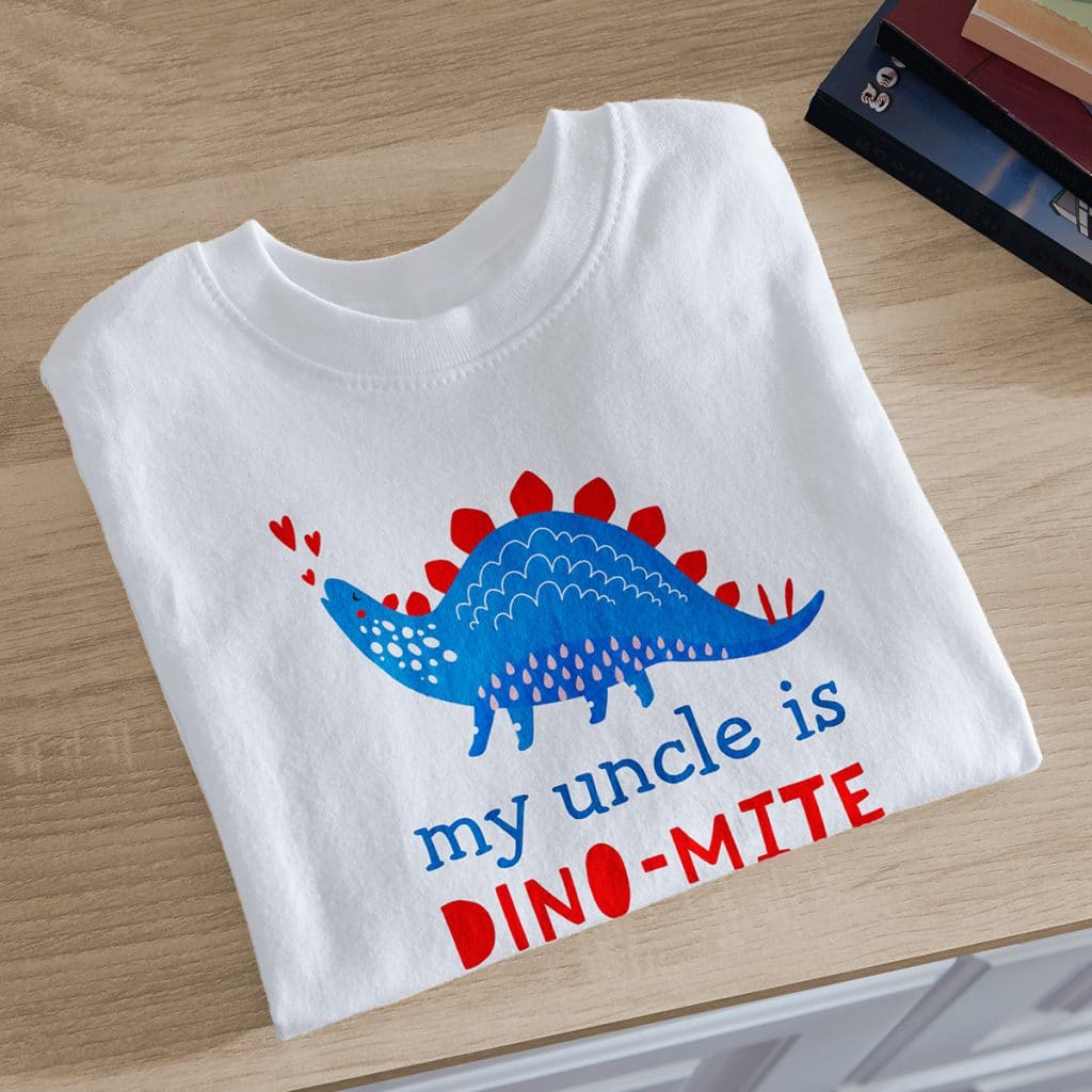 Image of a white toddler t-shirt, folded, bearing a design that reads "My uncle is Dino-Mite" and a dinosaur image.