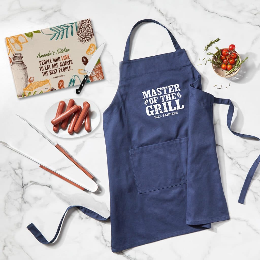 Flat lay image featuring navy blue apron with "Master of the Grill" design, cutting board, and other cook out essentials