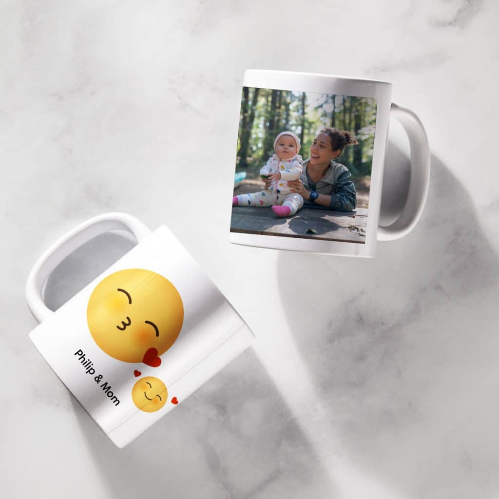 Flat lay image showing two white mugs — one with a kissing emoji design that reads "Philip & Mom" and one with a sweet photo of a mother and her baby daughter