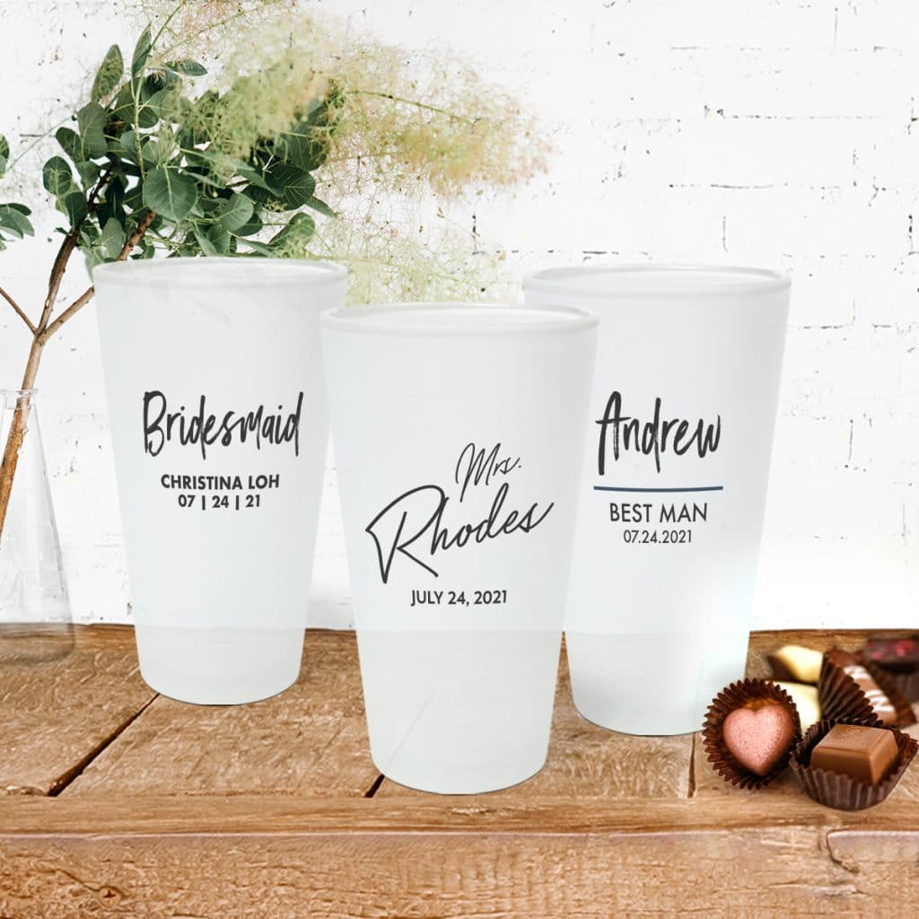 3 frosted pint glasses sitting on a table. Each pint glass features a different wedding-centric design.