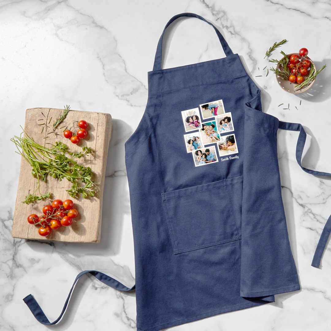 Image of navy apron with photo design laying on a countertop, surrounded by vegetables, herbs, and cooking materials