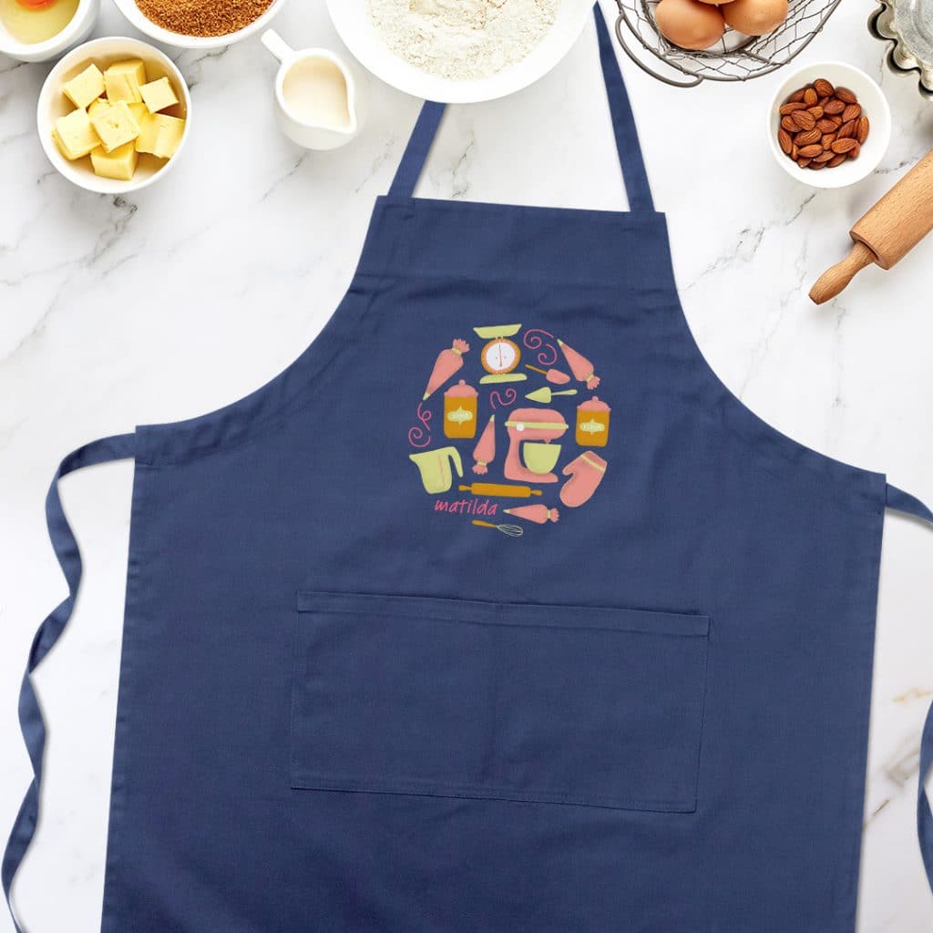 Flat lay image of a navy apron on a countertop. Apron is featuring a baking design and the name "Matilda".