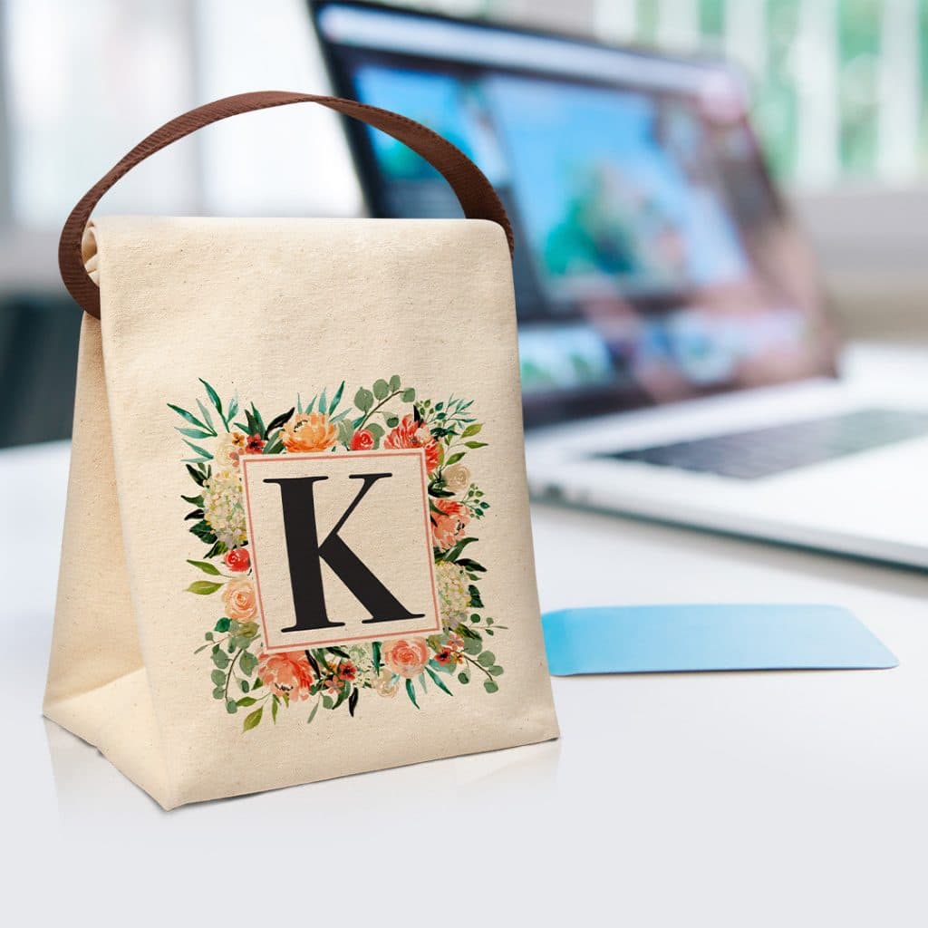 Image of a lunch bag with a large letter K surrounded by a floral frame. There's a laptop computer out of focus in the background.