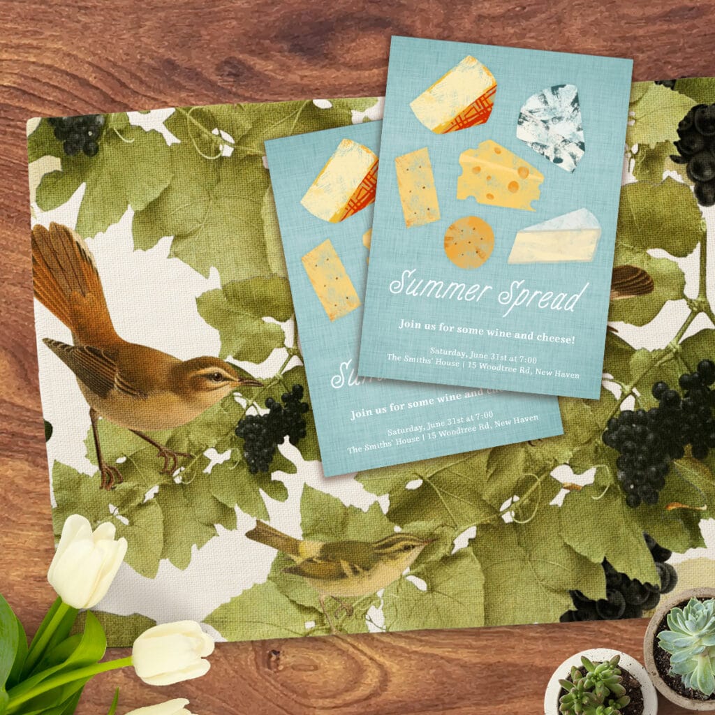 Flat lay image featuring cloth placemat with vineyard-inspired design. On top of the placemat is invitations for a wine and cheese party.