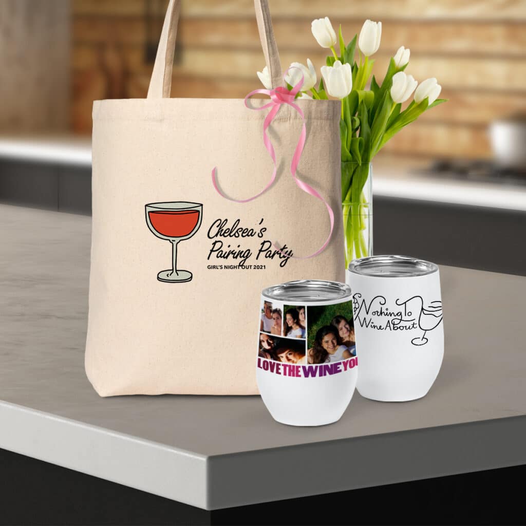Image of a canvas tote bag with "Chelsea's Pairing Party" design sitting on a countertop. In front of the bag are two insulated wine tumblers, both featuring wine-centric designs.