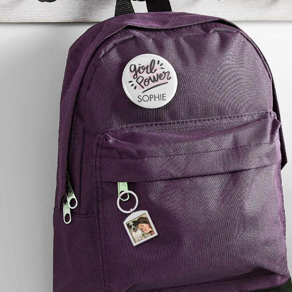 Image of a purple backpack with a custom button pin with a "Girl Power" design and the name Sophie. There's also a photo keychain hanging from the zipper.