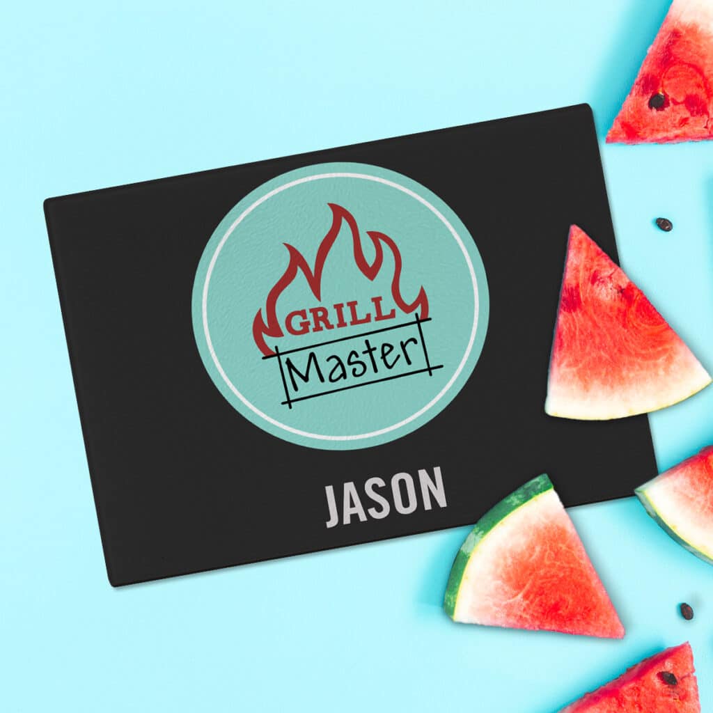 Flat lay image of watermelon slices on top of a cutting board. The glass cutting board features a design that says "Grill Master" and the name Jason.