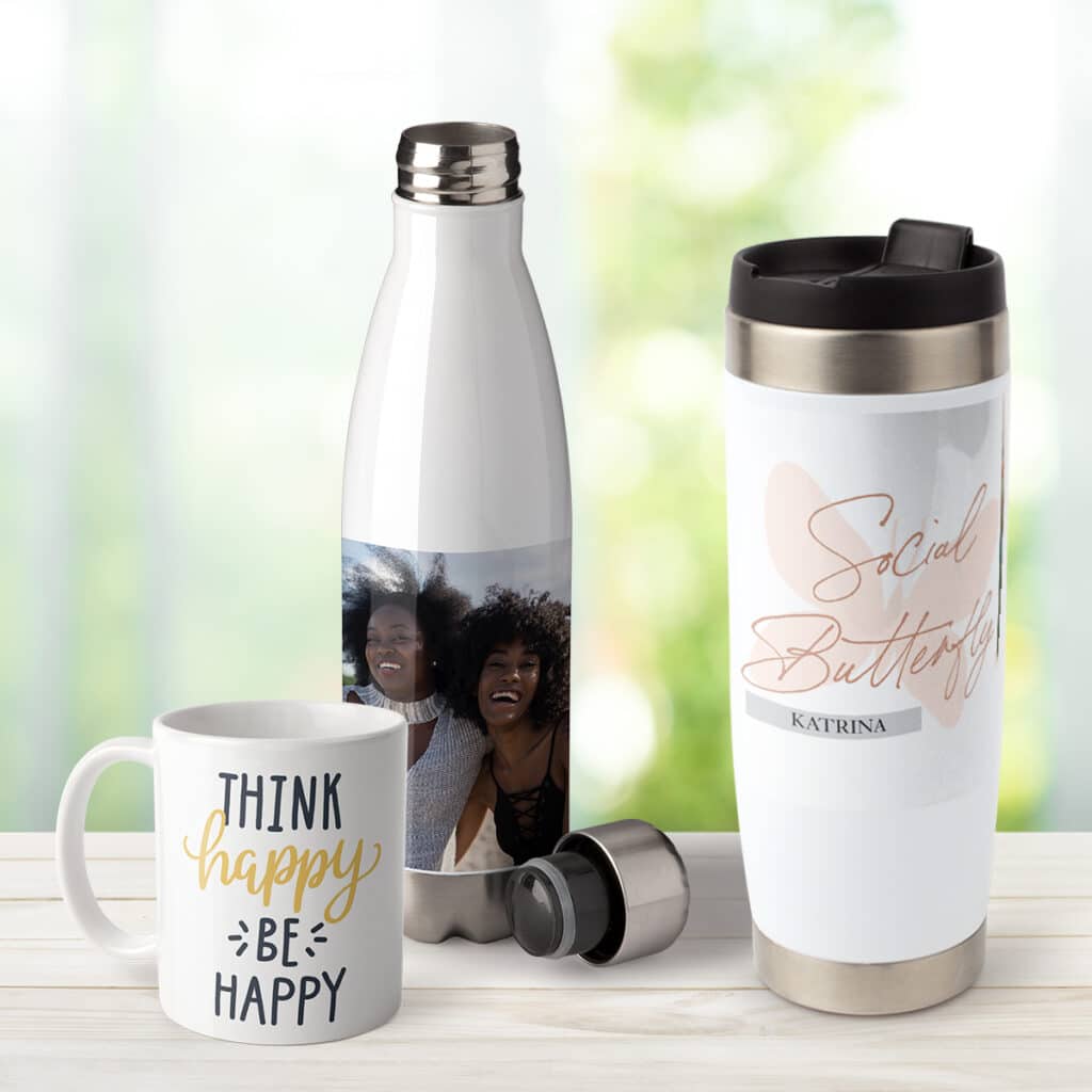 Image of a coffee mug, insulated water bottle, and travel tumbler with custom text and photo designs on a picnic table outdoors.