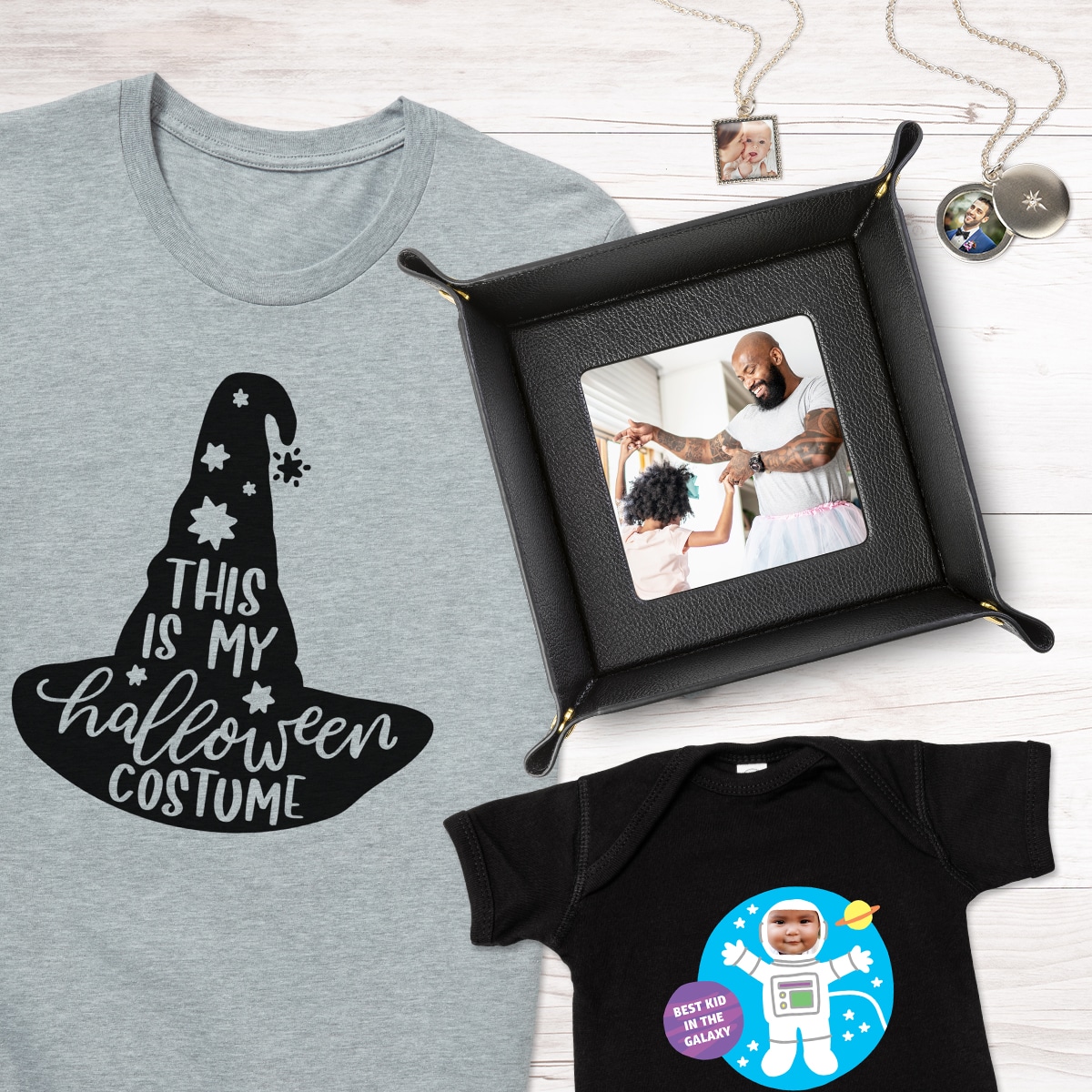 An adult t-shirt with a halloween design, a toddler t-shirt featuring an astronaut design, a valet tray featuring a photo of a dad and daughter dancing, and two photo necklaces are all laying on a tabletop.