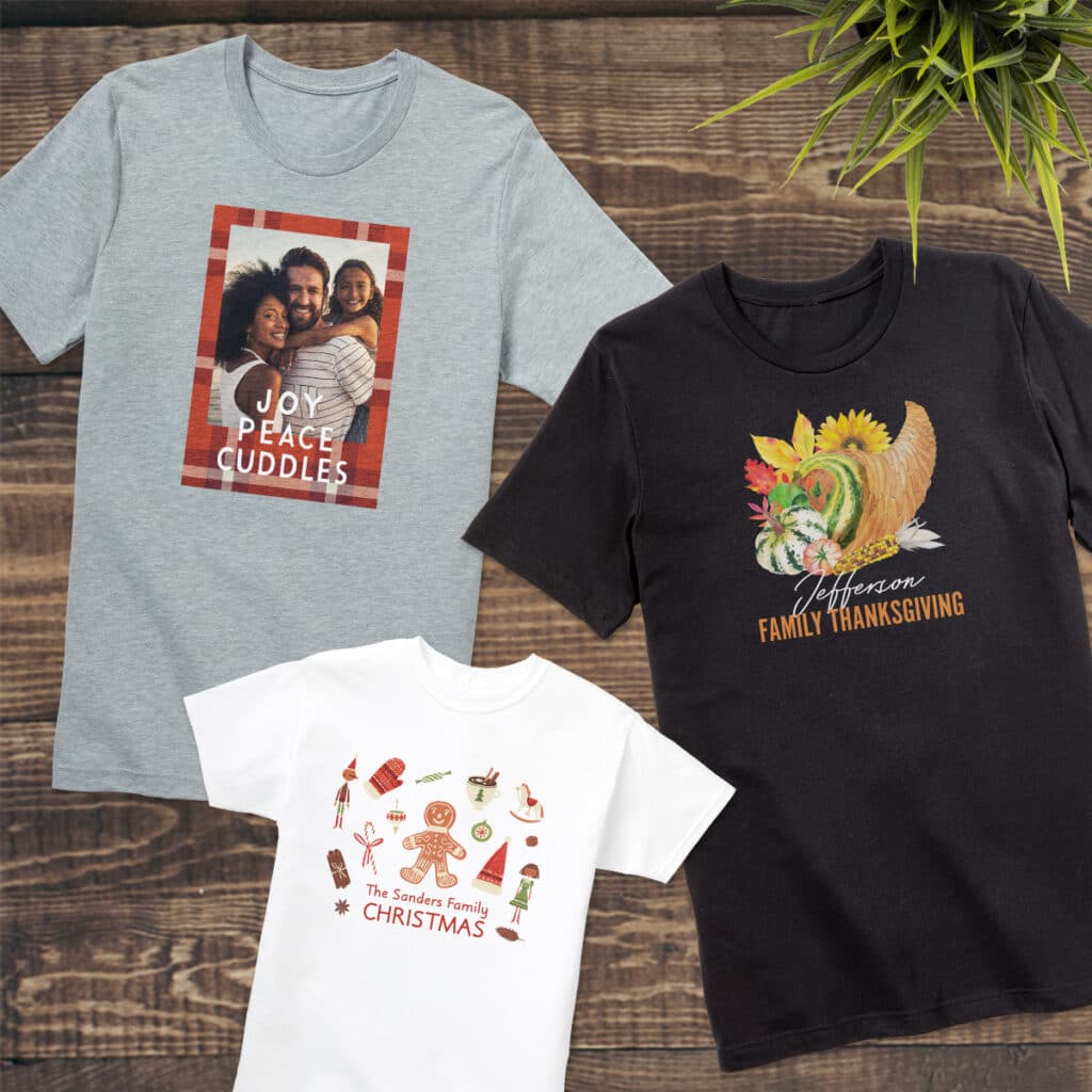 Three custom t-shirts laying on a tabletop, all with festive holiday designs for Christmas and Thanksgiving. Decorative foliage is laying beside them.