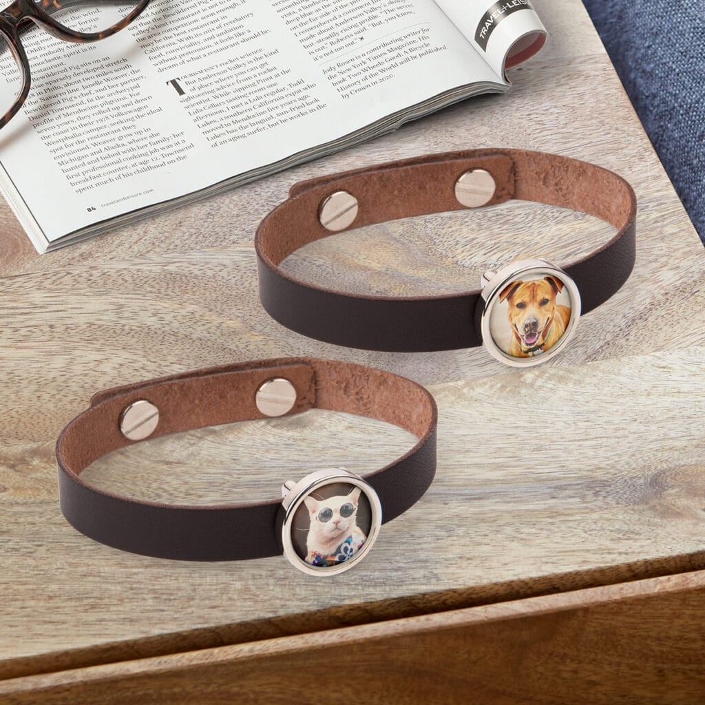 Two leather photo charm bracelets laying on a countertop, one featuring a photo of a cat and the other a dog. A magazine and pair of glasses sits behind them.