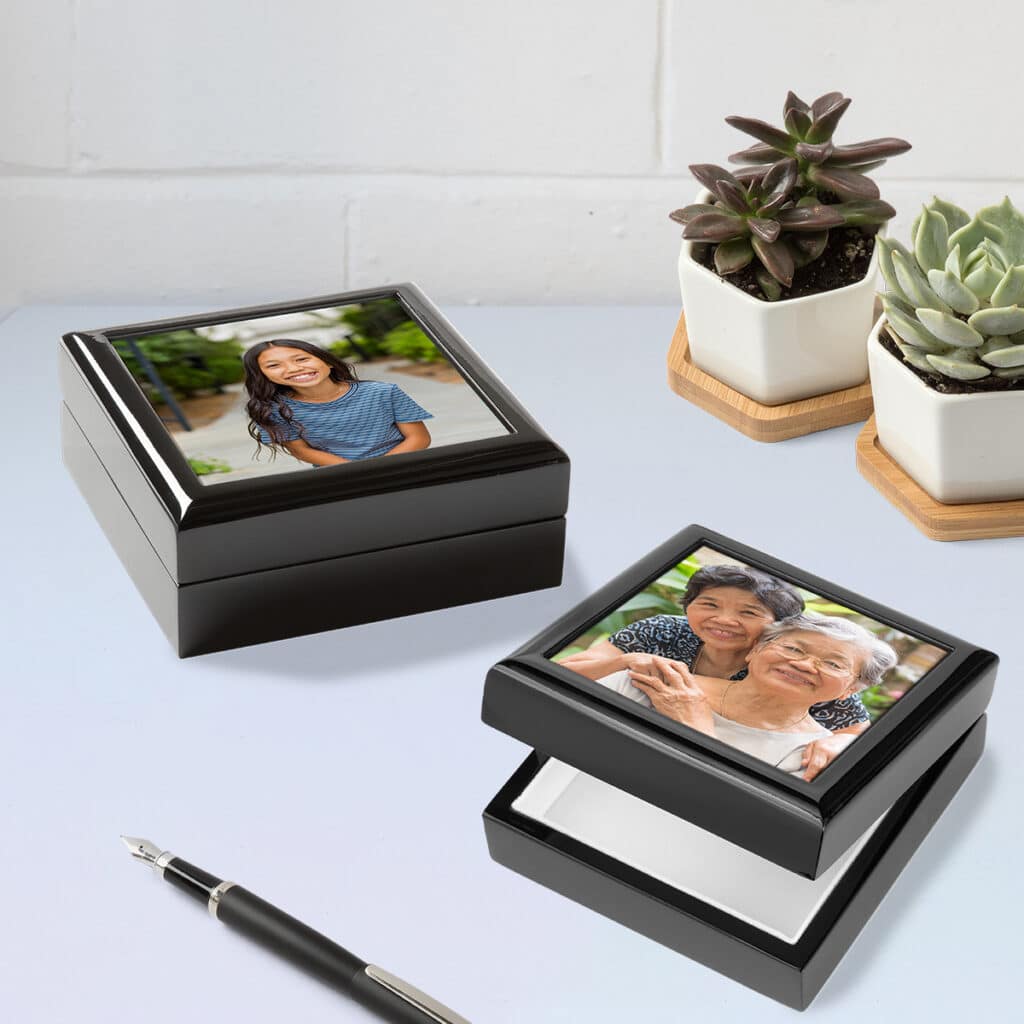 Two photo keepsake boxes laying on a countertop with photos featuring a young girl and a mother + grandmother. An ink pen lays in front of the boxes with two succulent plants behind them.